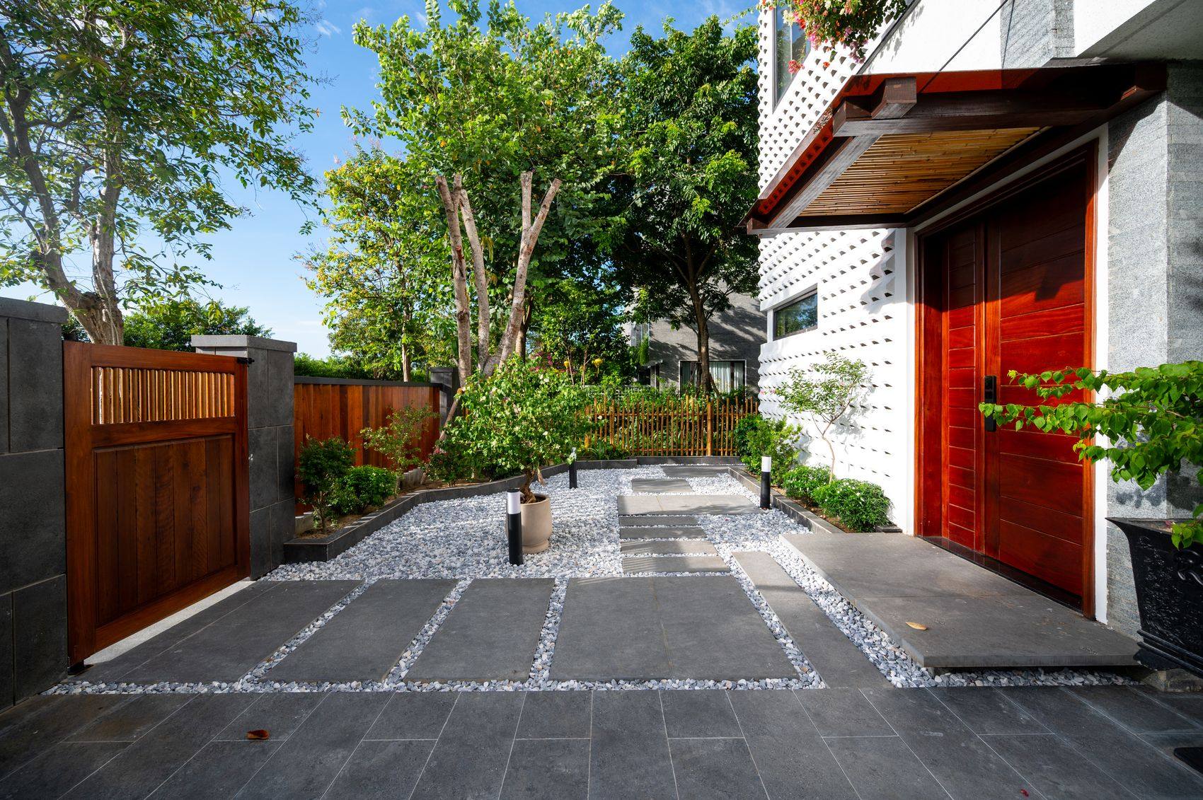 Lối vào NAMO Villa Hội An ban ngày — sân lát đá phiến xám kẻ ô sỏi trắng dẫn tới cửa gỗ nâu đỏ có mái hắt, tường gạch bông gió trắng bên phải, cổng gỗ và cây xanh bên trái