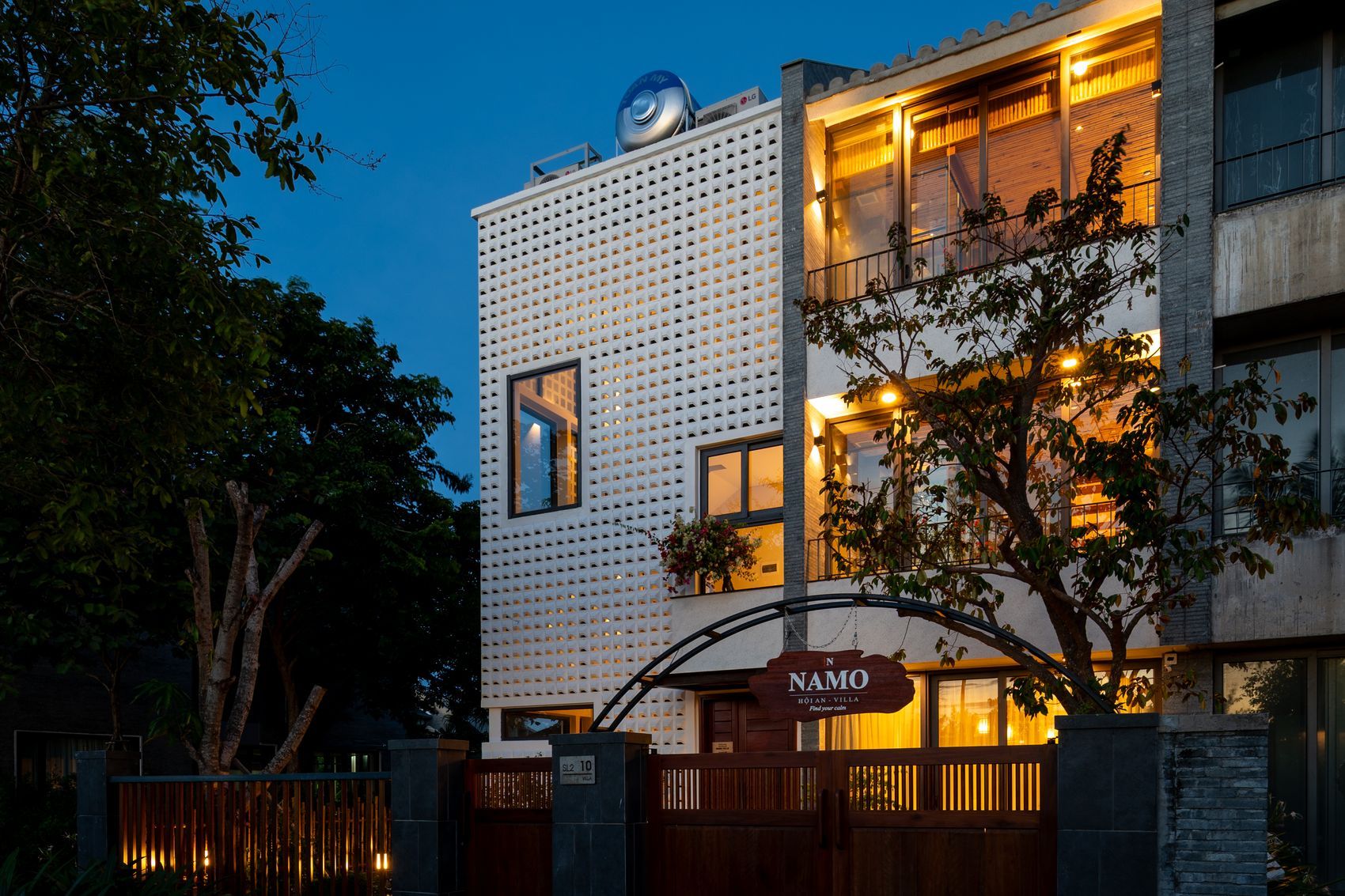 Mặt tiền NAMO Villa Hội An lúc lên đèn nhìn nghiêng từ đường — nhà ba tầng tường gạch bông gió trắng, các ô cửa sáng đèn vàng, biển gỗ NAMO dưới khung vòm sắt và cổng gỗ nâu đỏ