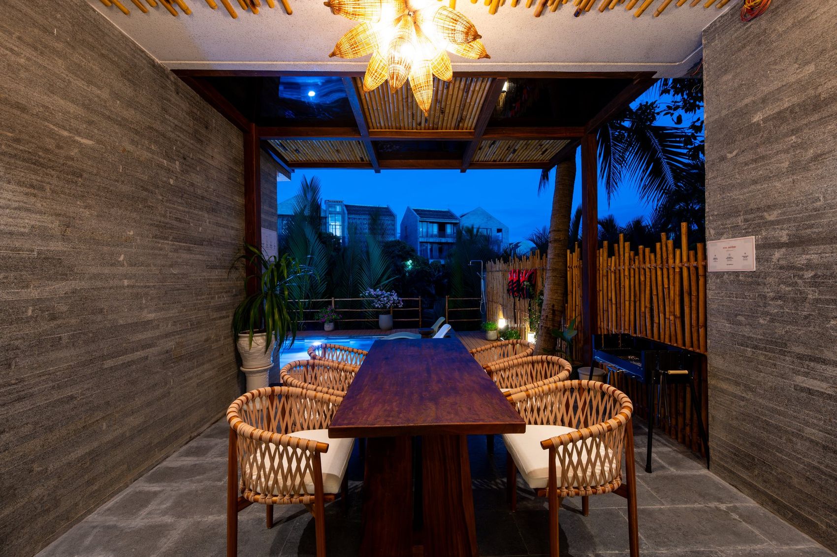 Khu pergola ngoài trời NAMO Villa Hội An lúc chạng vạng — bàn gỗ dài tám ghế mây, đèn mây hình hoa, bếp nướng than bên phải, nhìn xuyên ra hồ bơi
