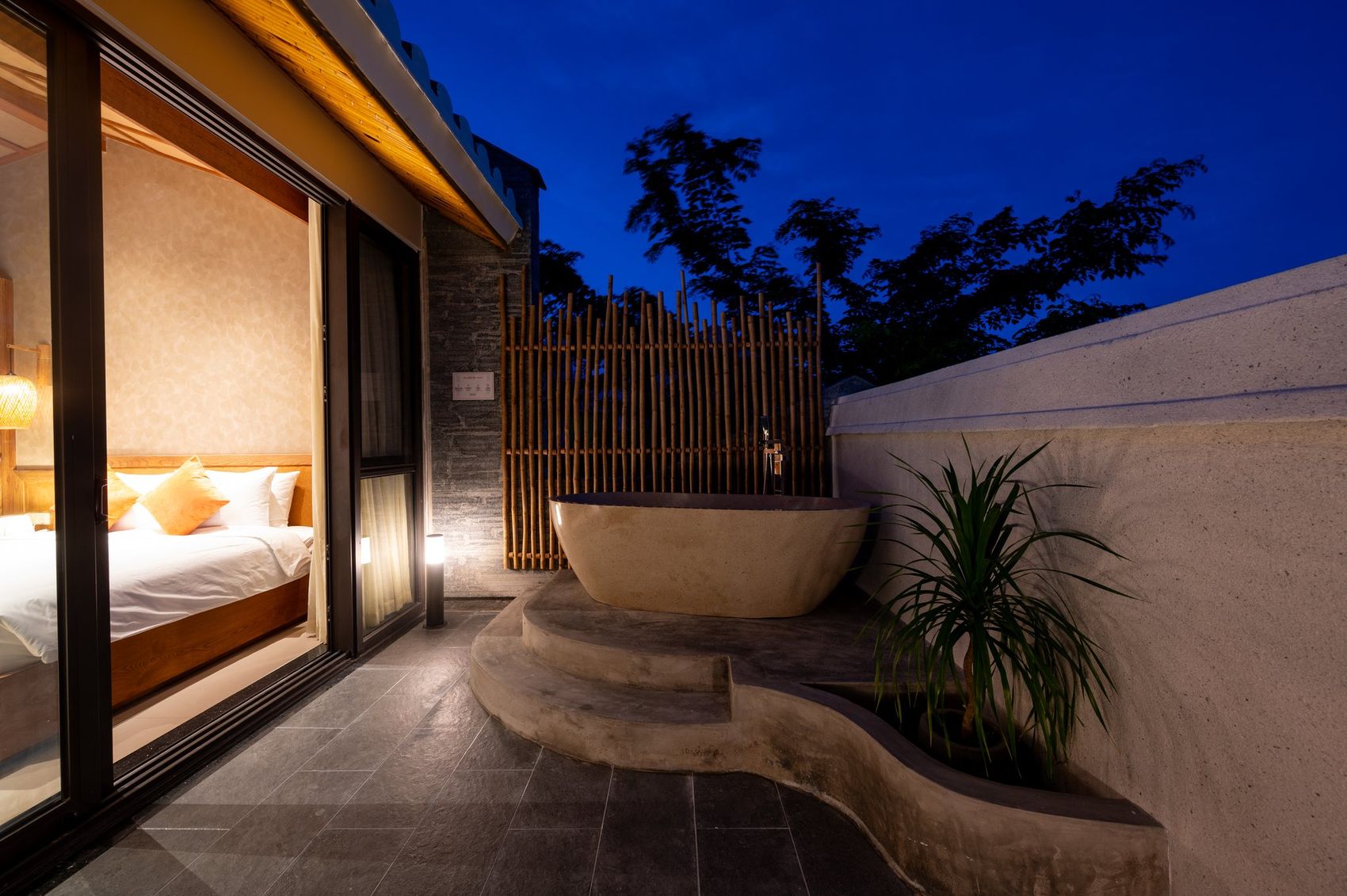 Sân thượng riêng của phòng Sky Soaking tại NAMO Villa Hội An lúc chạng vạng — bồn ngâm đá freestanding trên bệ, sen tắm đứng, vách tre, và cửa kính nhìn thẳng vào phòng ngủ sáng đèn ấm